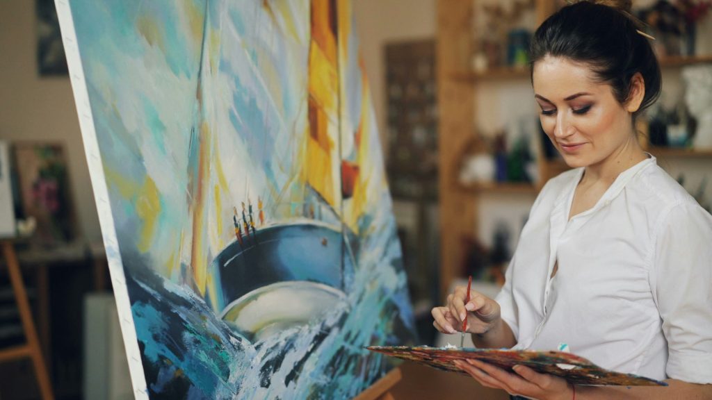 A woman paints a sailboat on a canvas.